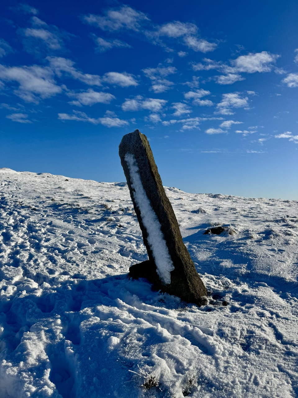 A snow-covered Long Stoop marker stone leans in its foundations; the backdrop is snowy landscapes and blue skies.