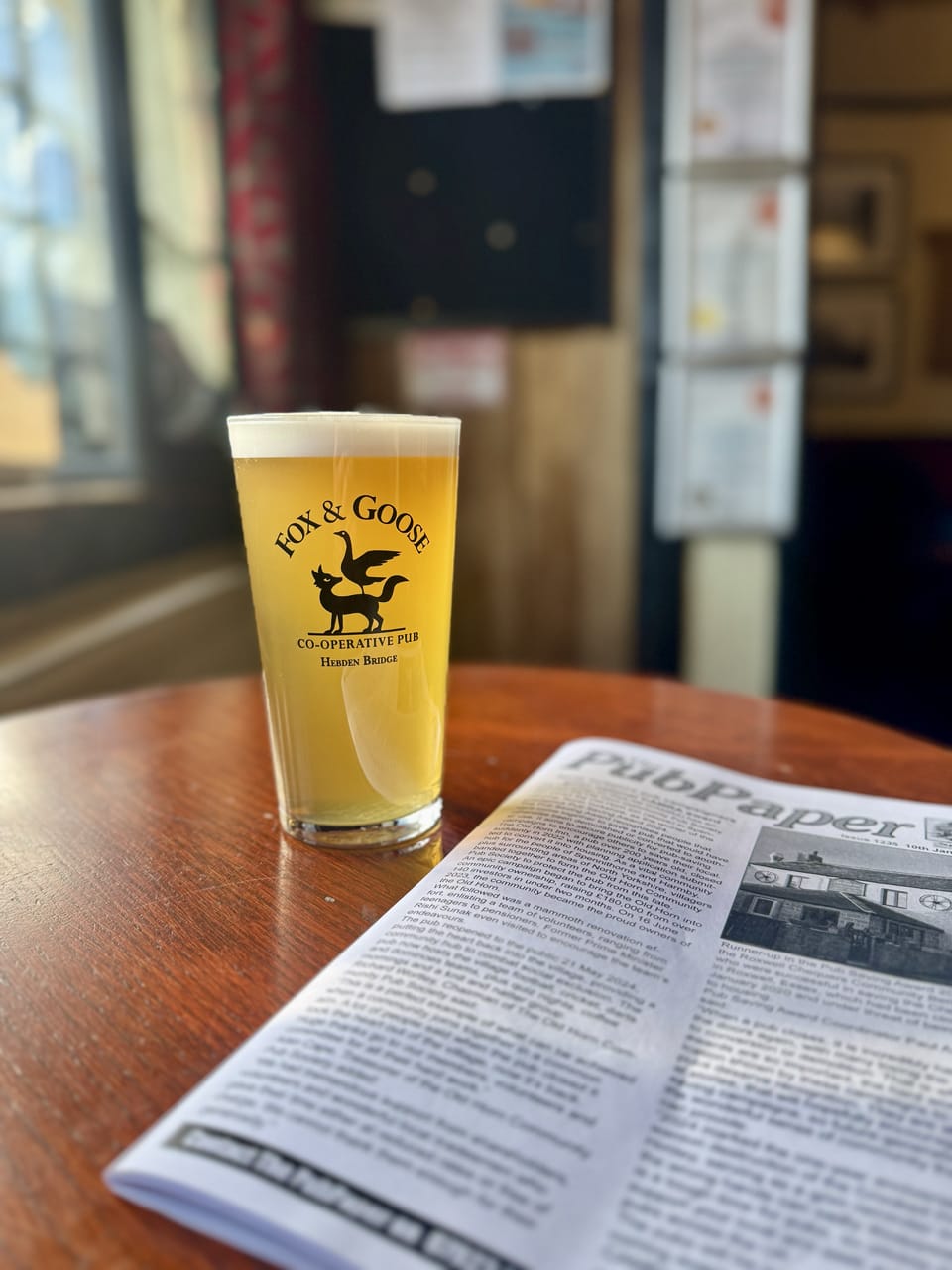 A golden pint of ale sits on a round wooden table next to a copy of The Pub Paper. It is illuminated by the winter sun.