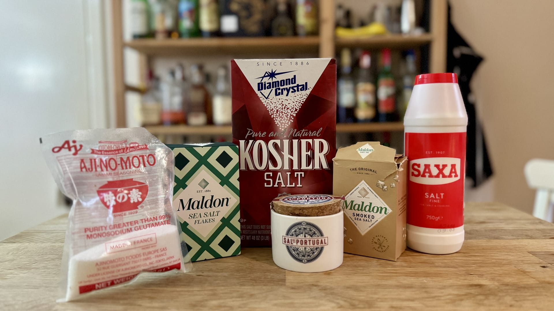 A landscape photograph of a lineup of different salts. From left to right: MSG, Maldon sea salt, Diamond Crystal kosher salt, Portugese fleur de sel, Maldon smoked sea salt, and SAXA table salt.